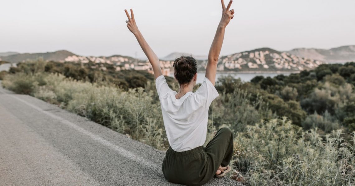 Woman on a roadside in Antalya raises peace sign, embracing summer with mountain views and tranquility.